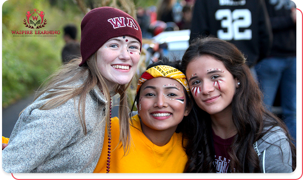 Close-up photo of three diverse female students smiling enthusiastically at a school event, likely a sporting game. They are wearing face paint and school gear, including a maroon beanie with 'WA' on it, symbolizing strong school spirit and community engagement.