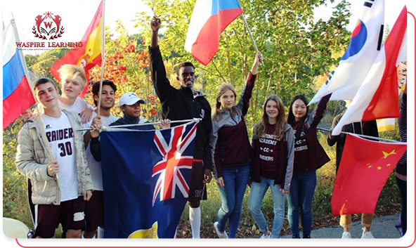 A diverse group of ten high school students, some wearing Washington Academy Raiders gear, standing together outdoors and enthusiastically holding up numerous international flags, including those from the United Kingdom, Russia, Spain, South Korea, and China, symbolizing global enrollment and community.